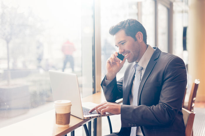 businessman in cafe talking on cell phone smiling at his lap top